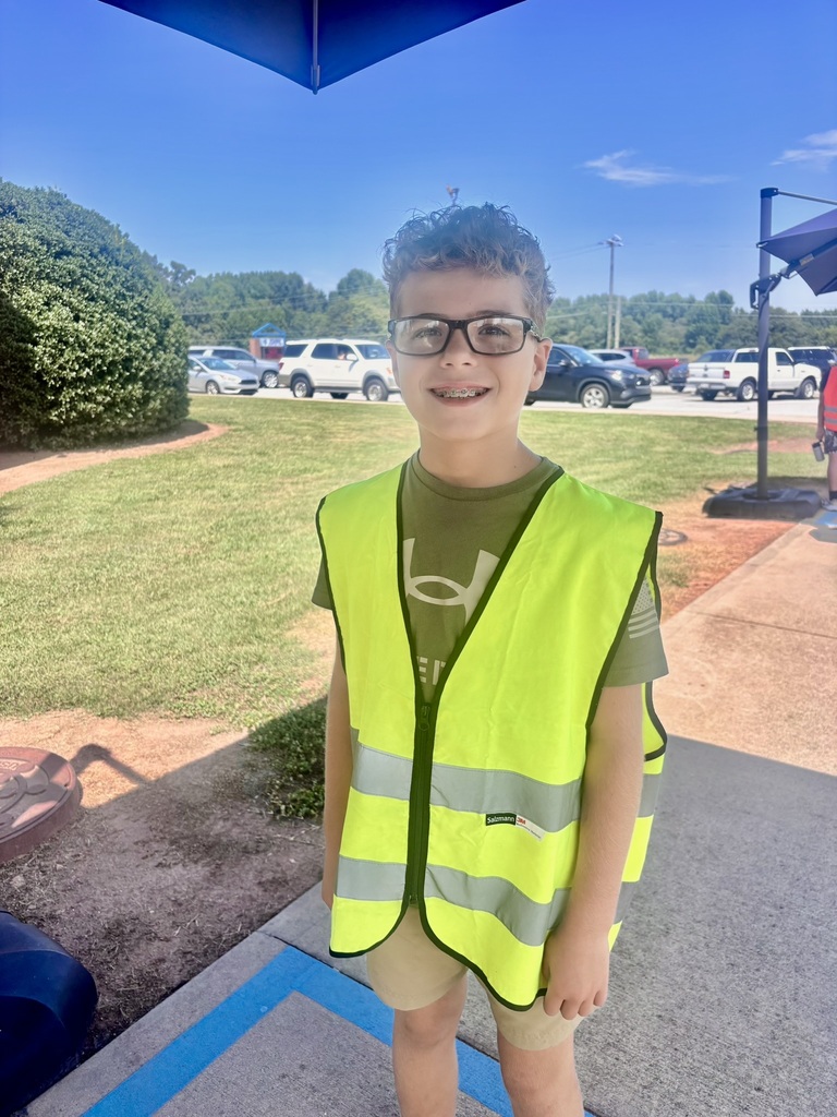 A smiling elementary student wearing glasses and a bright yellow safety vest stands outside the school near the car-rider area.