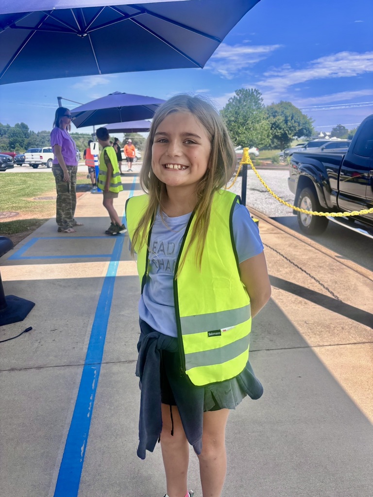 An elementary student wearing a bright yellow safety vest smiles while helping outside near the school car-rider line.