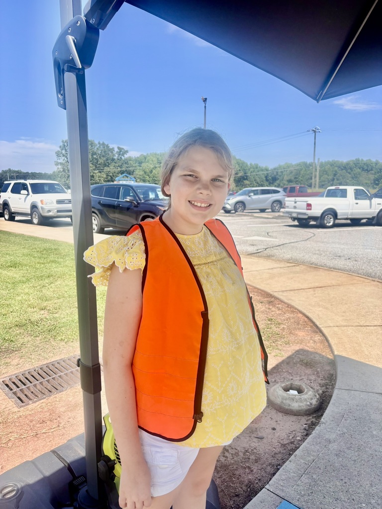 A smiling elementary student wearing a bright orange safety vest stands outside the school near the parking lot on a sunny day.