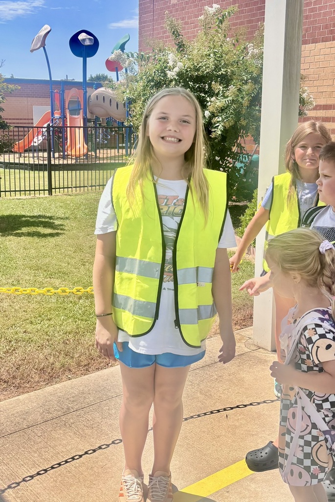 A smiling elementary student wearing a bright yellow safety vest stands outside the school near the playground as other students gather nearby.