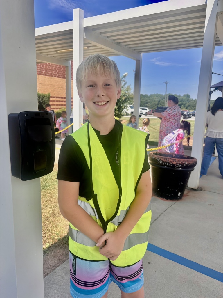 A smiling elementary student wearing a bright yellow safety vest stands near the school entrance as other students arrive in the background.