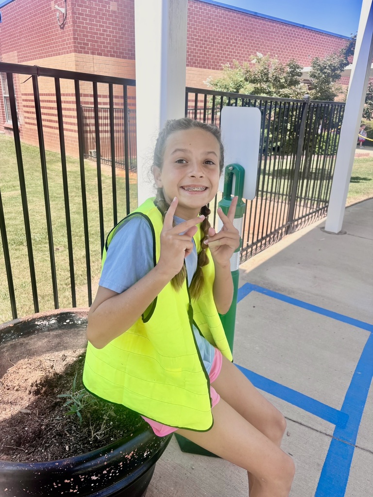 An elementary student wearing a bright yellow safety vest smiles and holds up peace signs while sitting outside the school.