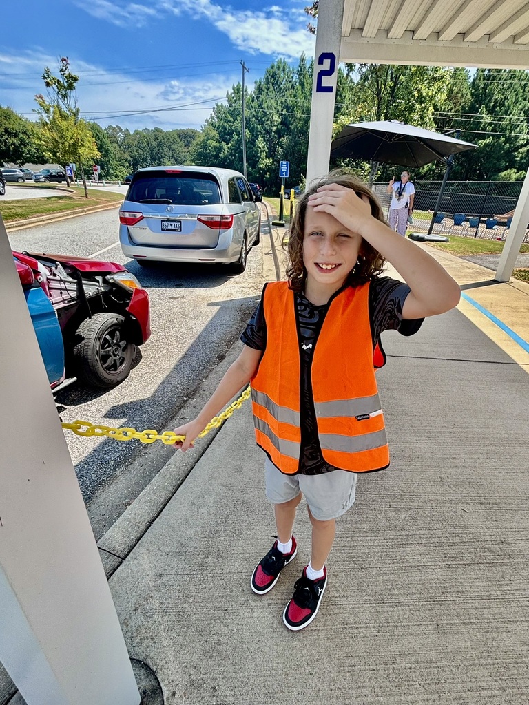 n elementary student wearing a bright orange safety vest smiles while standing near the school car-rider line on a sunny day.