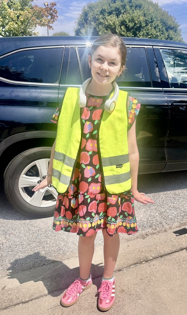 An elementary student wearing a bright yellow safety vest smiles outside the school while standing in front of a parked vehicle.