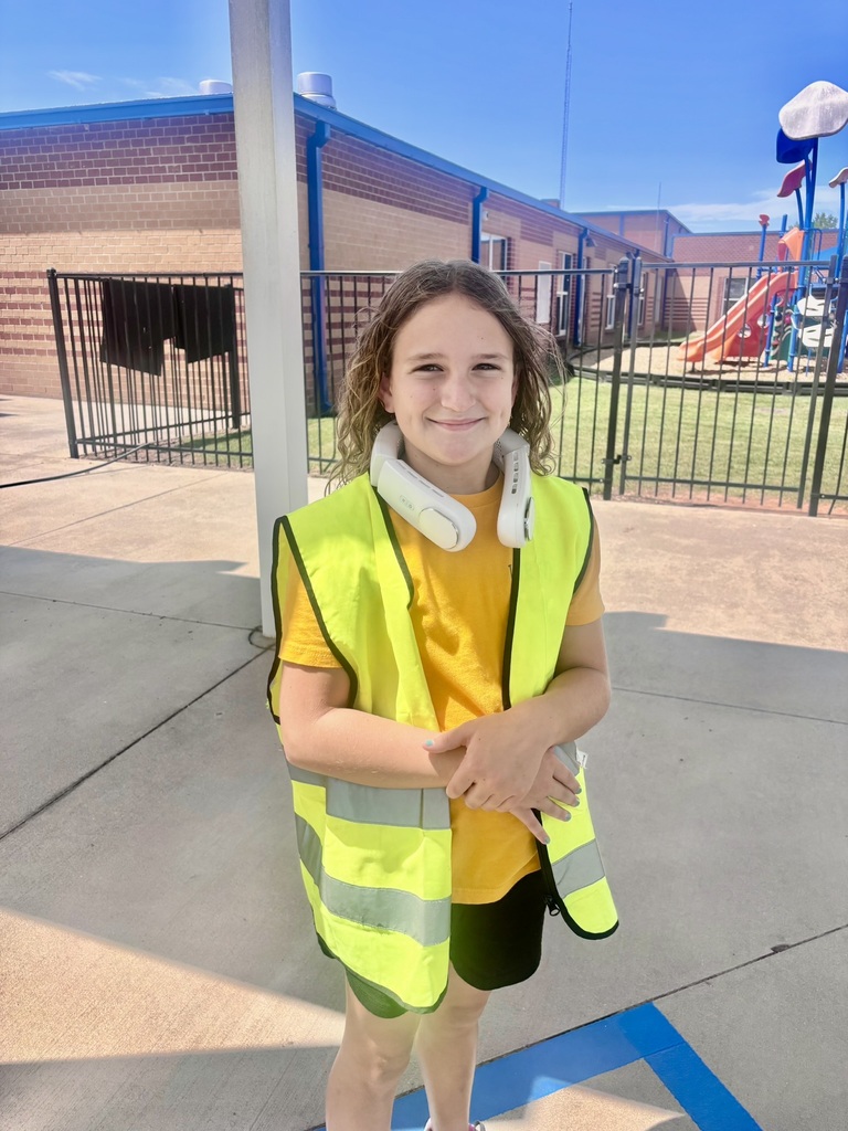 A smiling elementary student wearing a bright yellow safety vest stands outside the school near the playground.