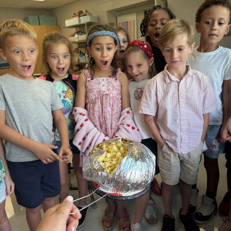Elementary students gather around with excited and surprised expressions as they watch popcorn being made in a foil-covered pan during a classroom activity.