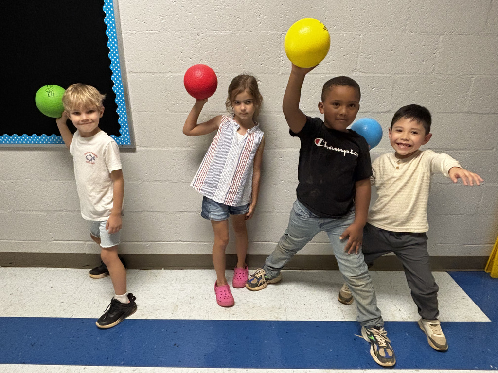 Four elementary students smile and pose with colorful playground balls during a physical education activity.