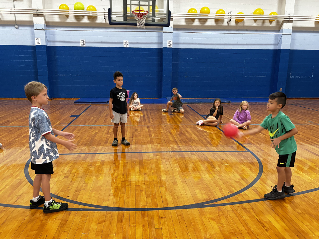 Elementary students participate in a PE activity in the gym as two students practice tossing and catching a red ball while classmates sit nearby waiting their turn.