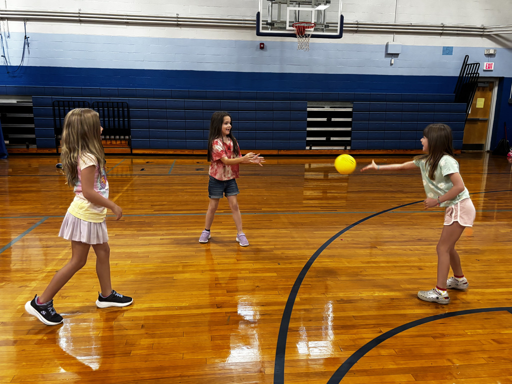 Three elementary students practice tossing and catching a yellow ball together during a physical education activity in the school gym.