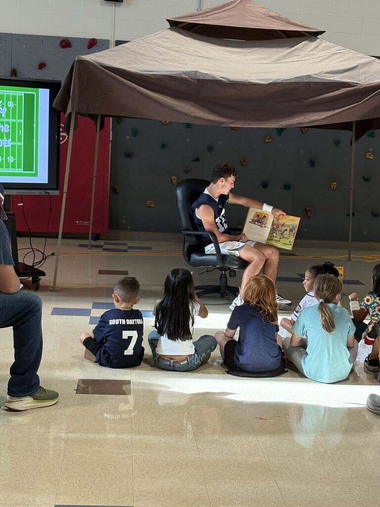 A student reads a book to a group of students in the school gym