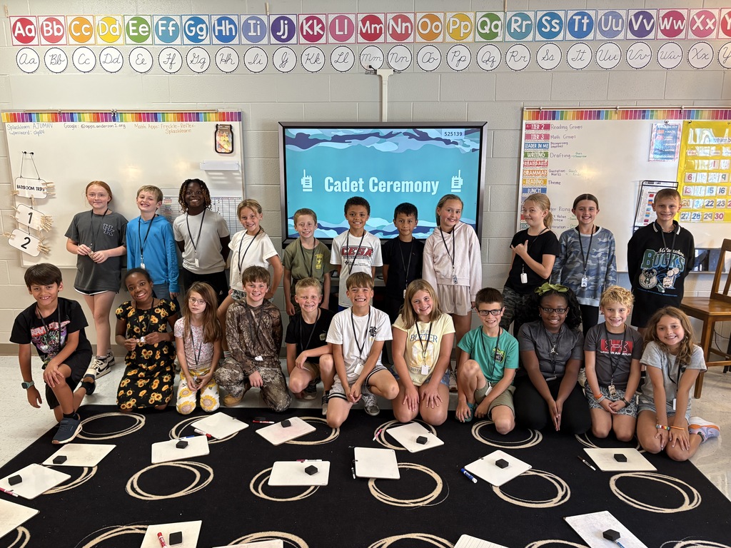 A group of elementary students pose together in a classroom after participating in "boot camp".  Students are gathered in two rows in front of an interactive display,  with classroom whiteboards and student work visible in the background. 