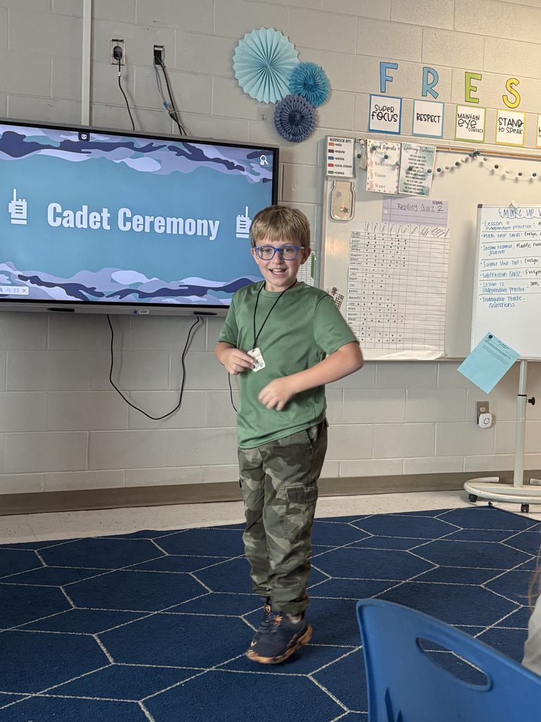 An elementary student wearing glasses and a green shirt smiles while standing at the front of the classroom during a Cadet Ceremony.  A classroom display behind him reads "Cadet Ceremony" with whiteboards and learning materials visible nearby.