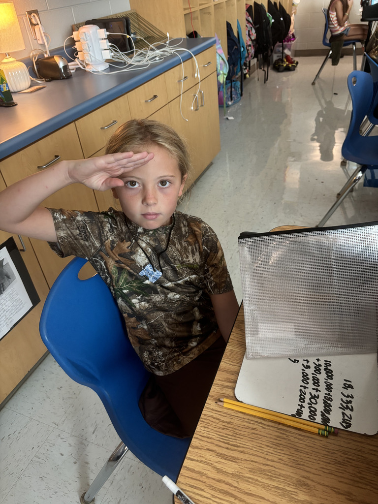 Student playfully salutes the camera while working on a math problem at her desk.