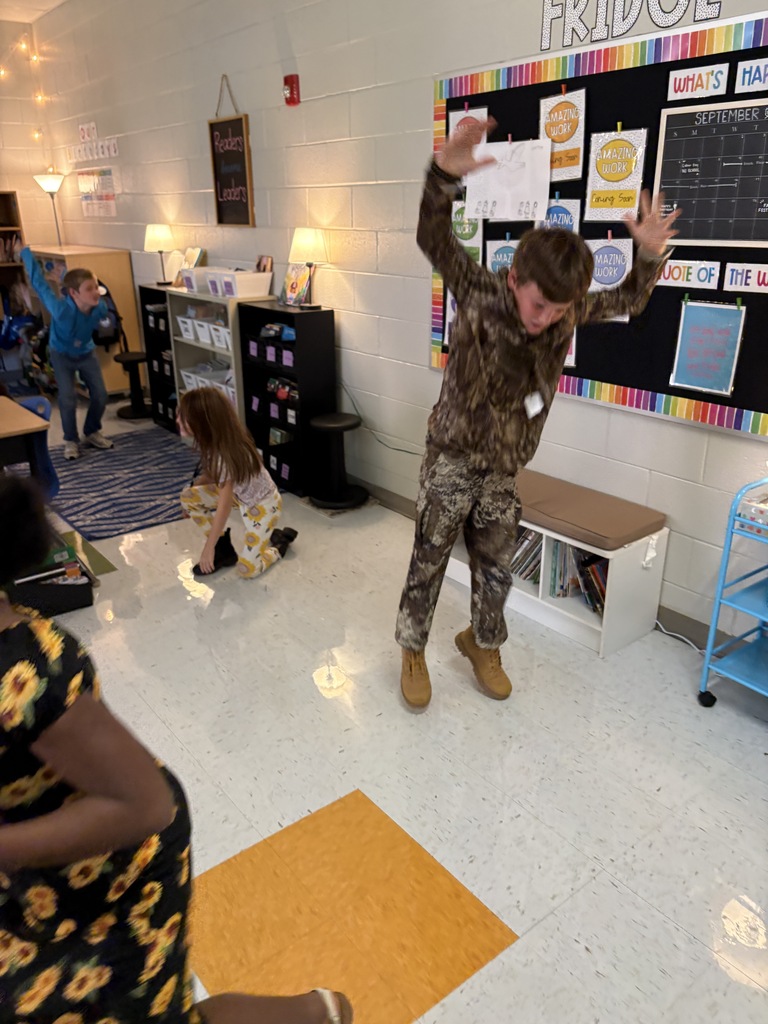 Elementary students participate in an interactive classroom activity, with one student in camouflage raising his arms while classmates move and play nearby.  Colorful classroom displays, bookshelves, and learning materials are visible in the background.