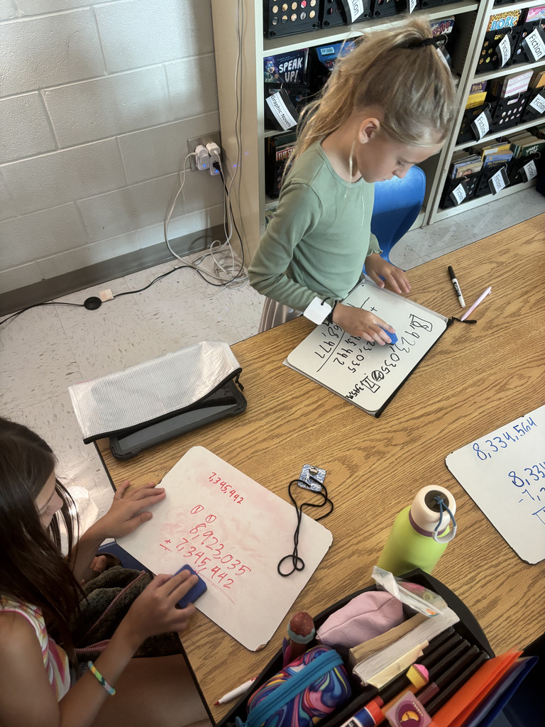 Two students practice math problems on dry-erase boards at a classroom table.