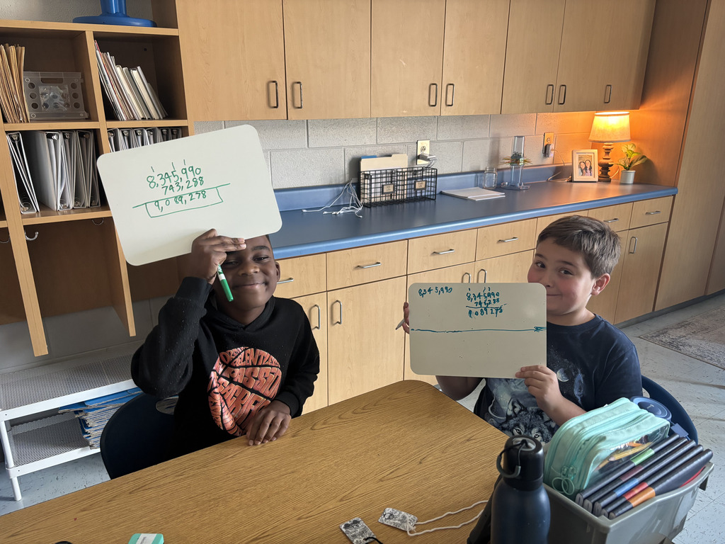 Two smiling students proudly hold up dry-erase boards showing their math work.