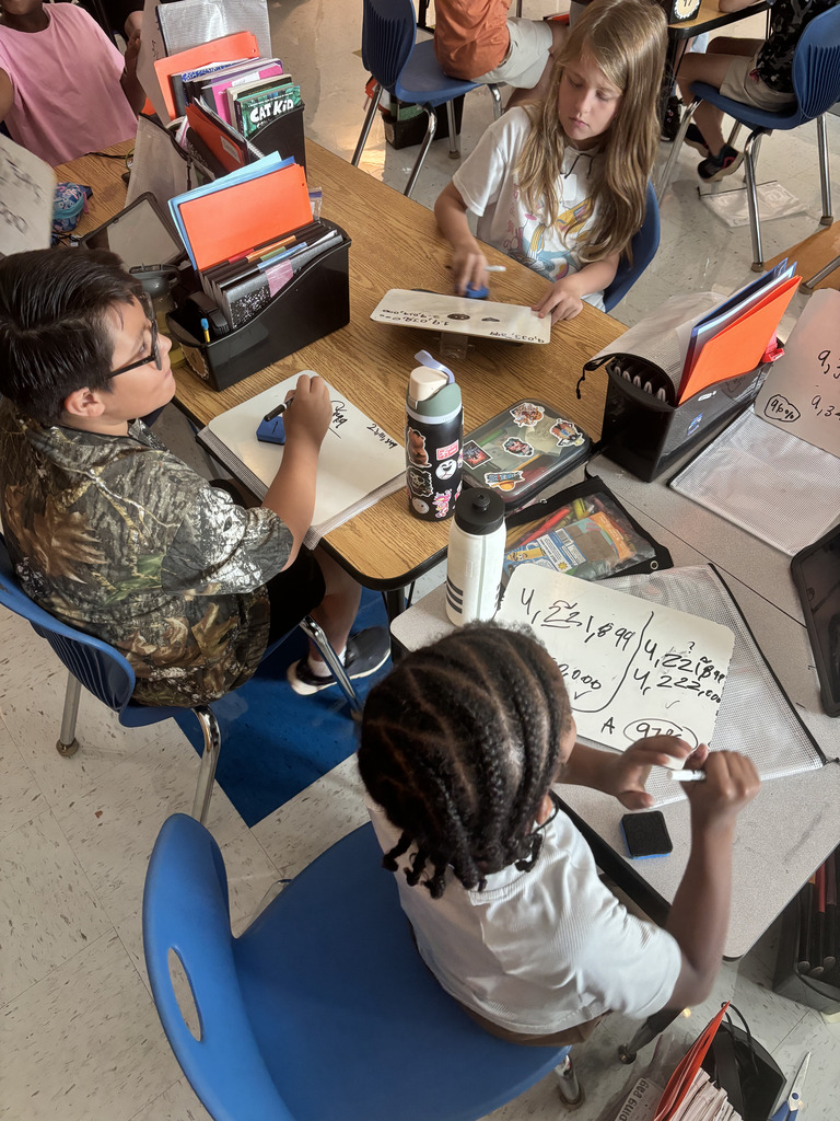 Two students practice math problems on dry-erase boards at a classroom table.