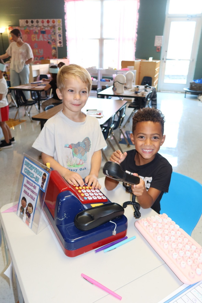 Two students smile while pretending to work at a reception desk during a classroom community helpers activity.