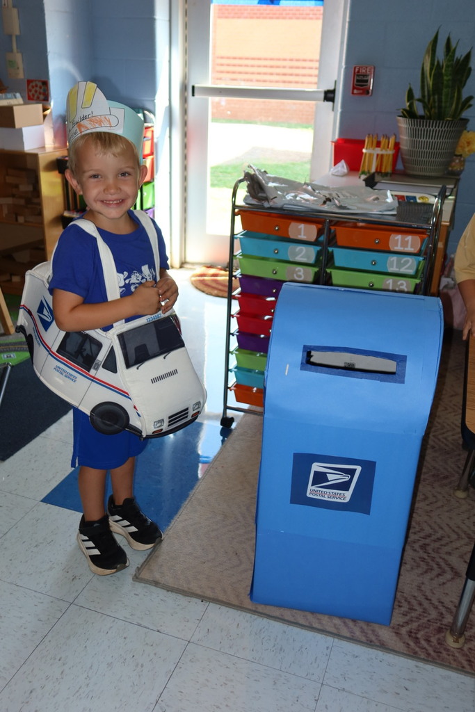 A student dressed as a mail carrier smiles beside a classroom mailbox during a community helpers activity.