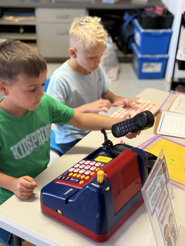 Two students pretend to work at a reception desk, using a phone and keyboard during community helpers day.