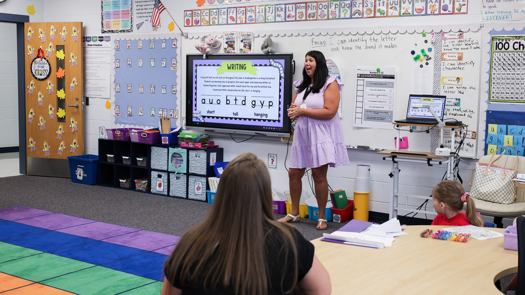 A teacher presents at the front of the classroom as students and a family member listen at a table