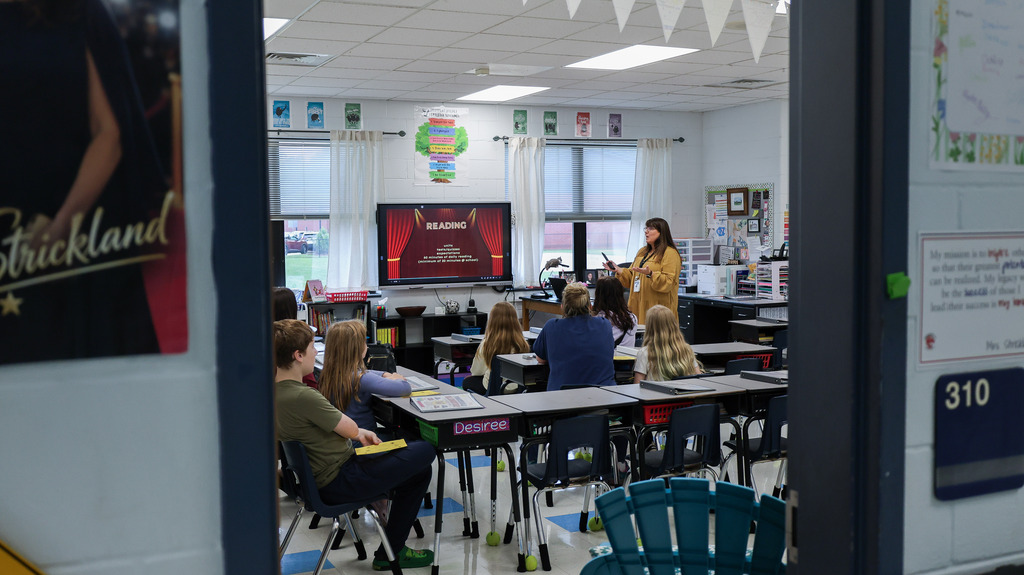 A teacher talks to students and parents in her classroom from the perspective of someone in the hallway