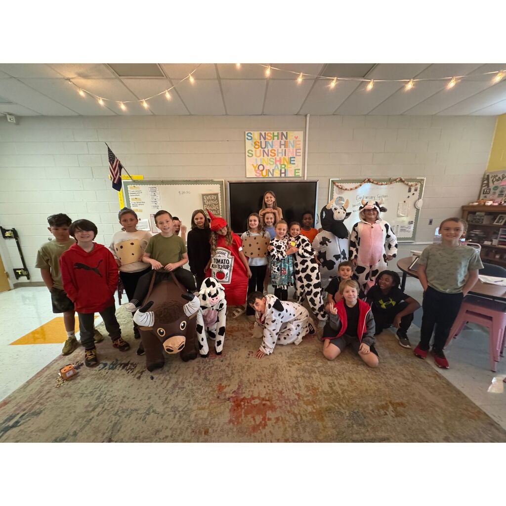 Students gather in a classroom wearing costumes inspired by foods and farm animals, including ketchup, cookies, and cows.