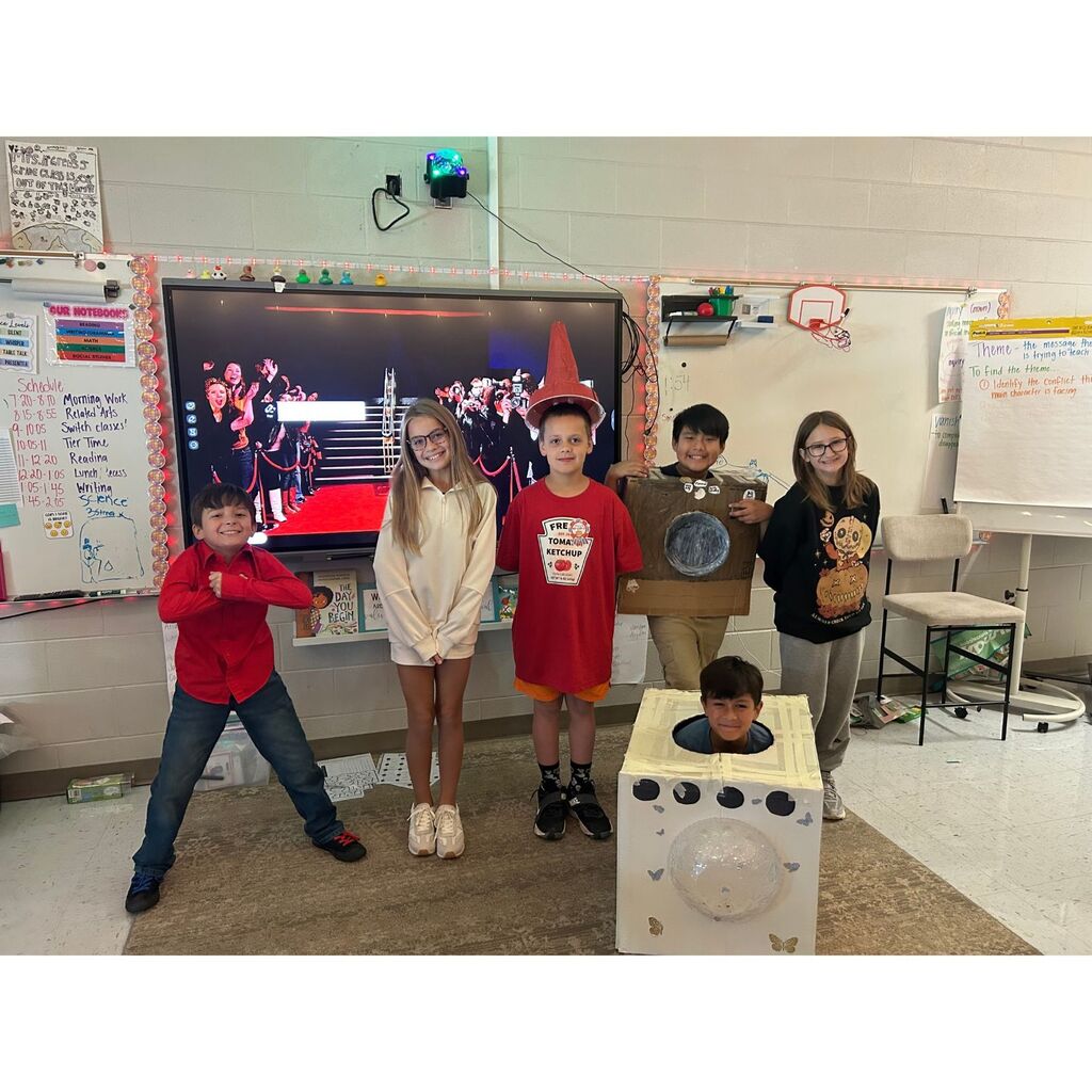 Six students pose in a classroom wearing creative costumes, including ketchup, a cardboard box, and a washing machine.