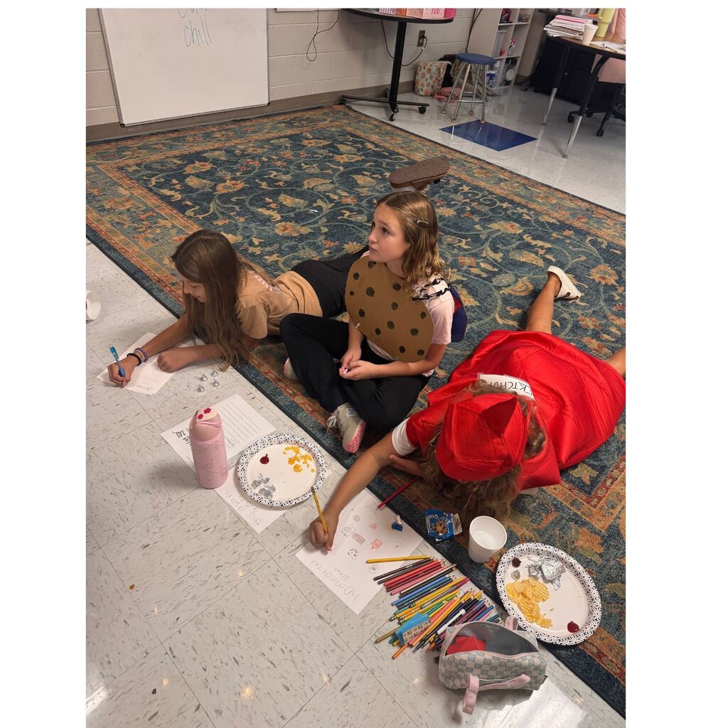 Three students in food-themed costumes sit and lie on the classroom floor while completing drawing and writing activities.