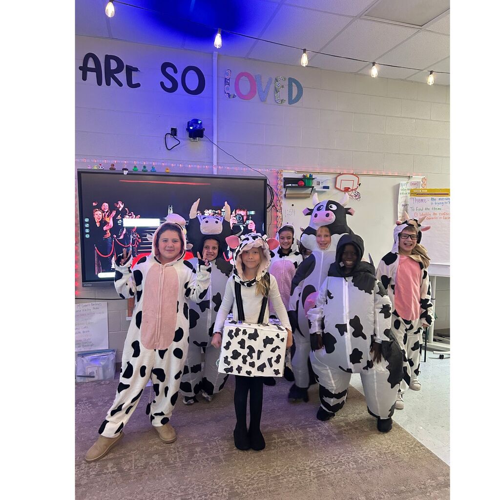 A group of students poses in a classroom wearing black-and-white cow costumes, including inflatable cow suits and homemade outfits.