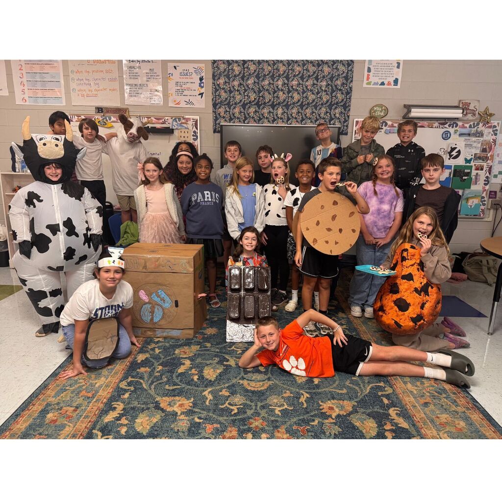 Elementary students pose together in a classroom wearing creative food-themed costumes, including cookies, chocolate, and a cow.