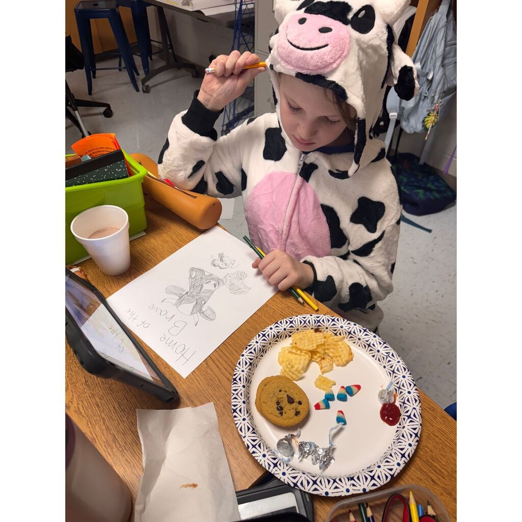 A student wearing a cow-print onesie sits at a desk drawing a cow beside a plate of snacks.