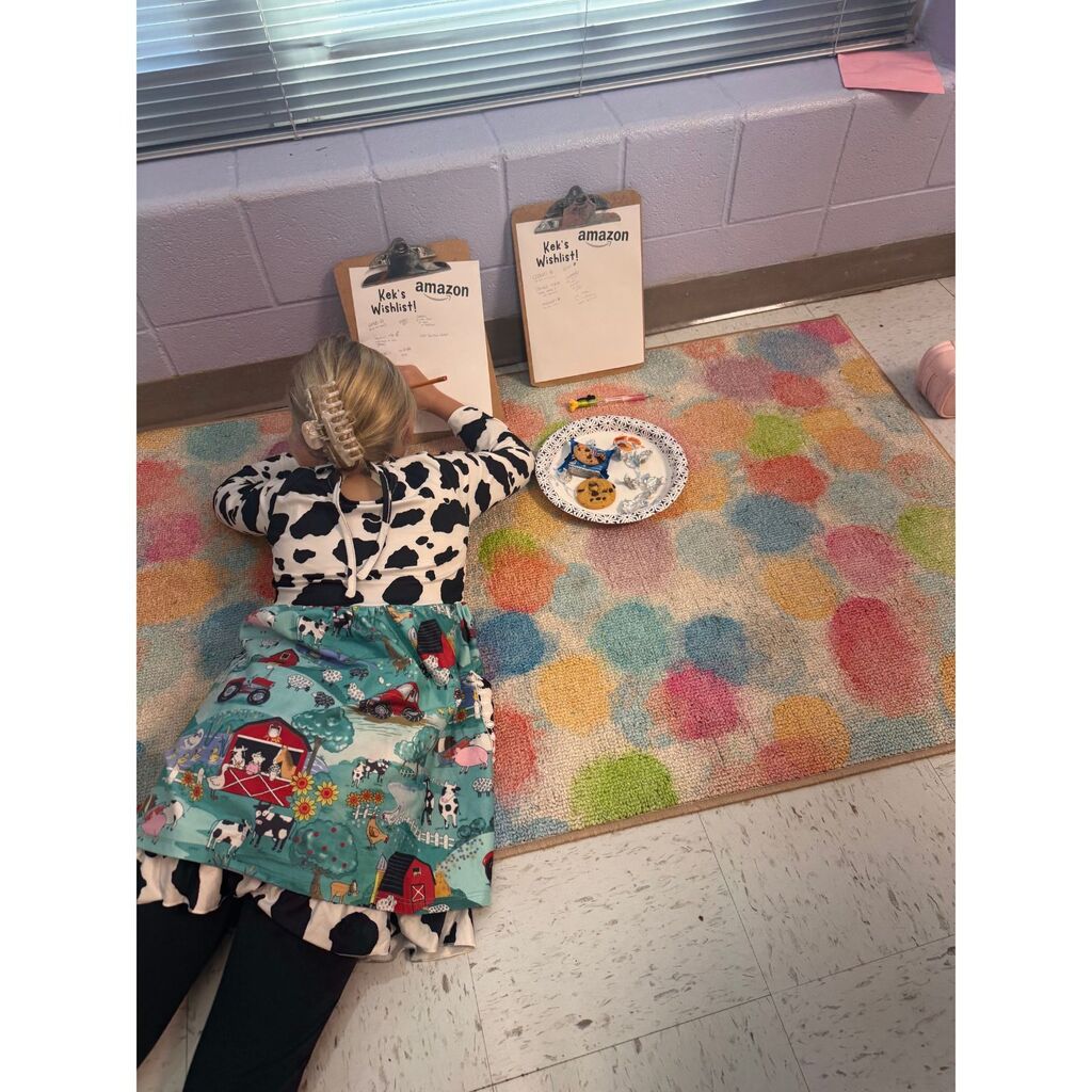 A student dressed in cow-print clothing lies on a colorful classroom rug while writing on a clipboard beside a plate of snacks.