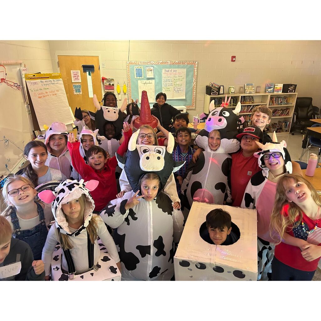 Elementary students pose together in a classroom wearing cow costumes and creative homemade cow-themed outfits.