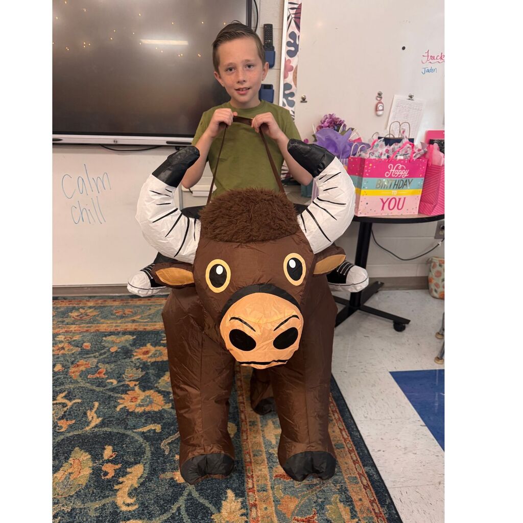 A student stands in a classroom wearing an inflatable brown bull costume with large white horns.