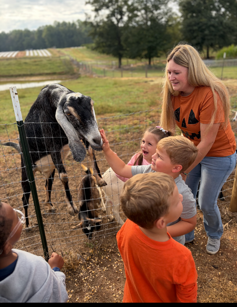3 k4 students petting the goat with a parent standing there smiling