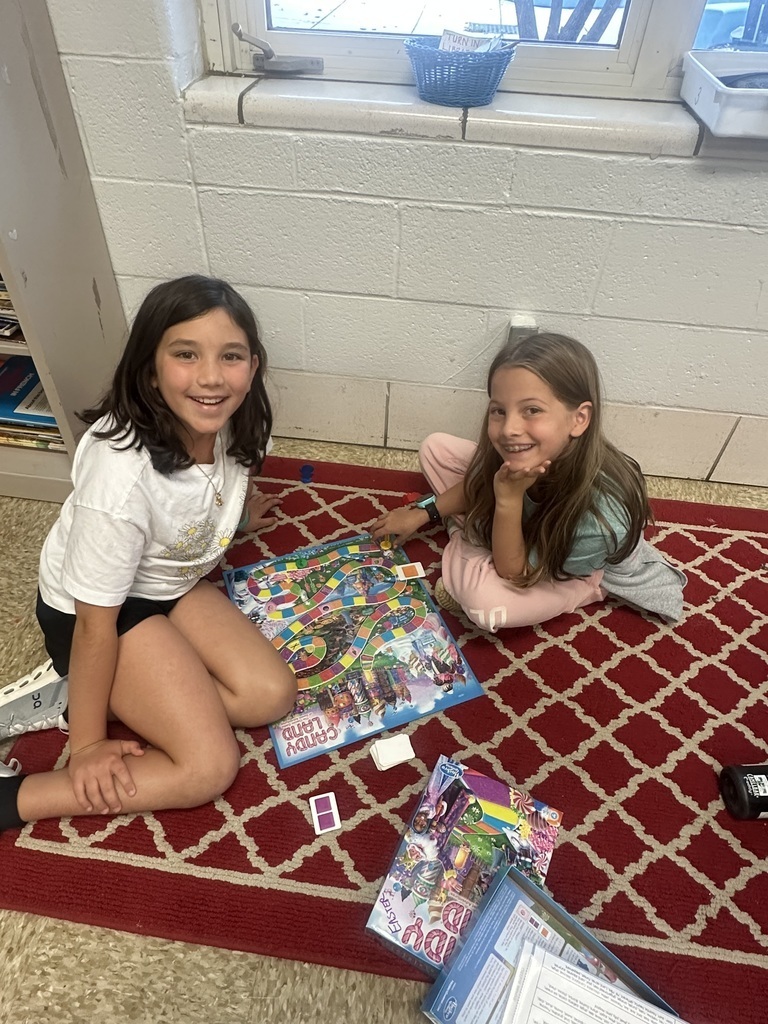 Two girls on a rug smiling with a board game. 