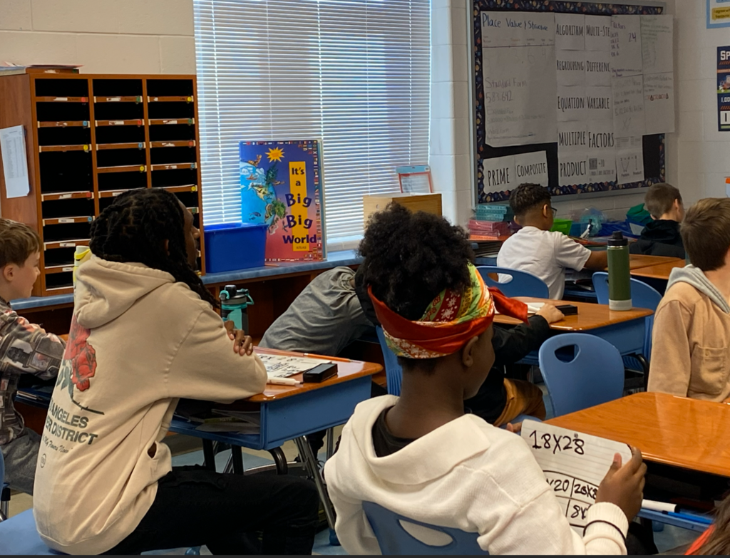 the teacher is sitting at a student desk as the student leads the class