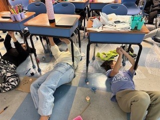 Students laying under their desks to paint artwork that is above them. 