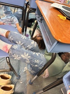 Students laying under their desks to paint artwork that is above them. 