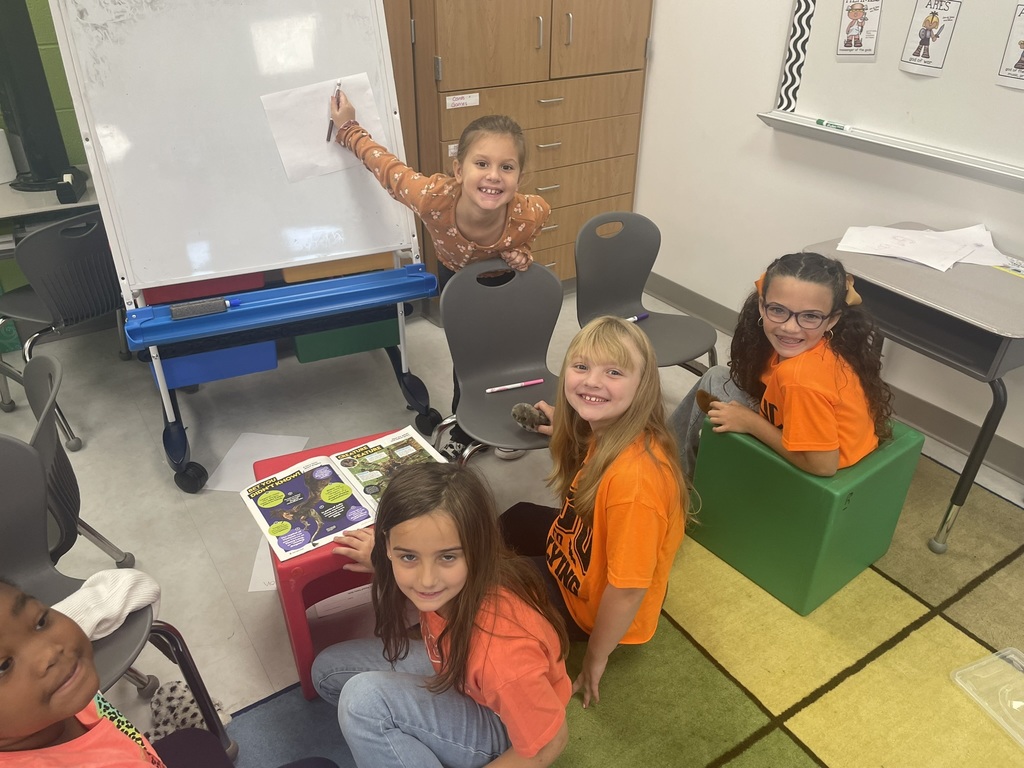 Students on carpet, wearing orange and facing a white board lesson