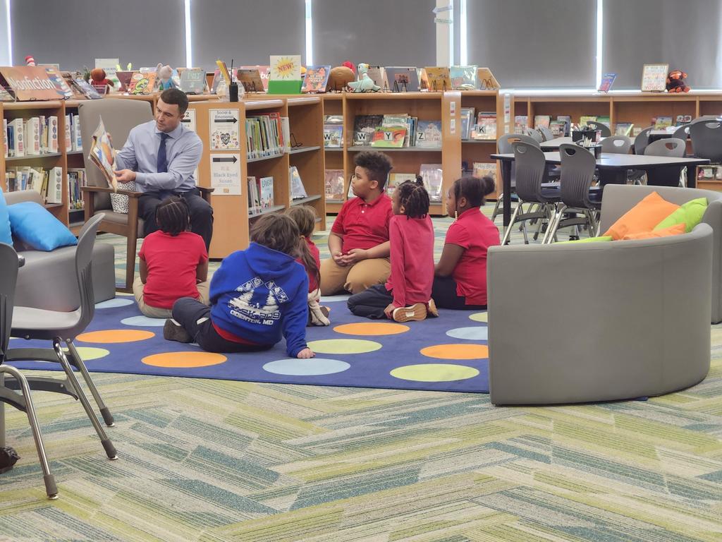 Photo of Principal Haslun reading a book to students in media class, sitting on a carpet.
