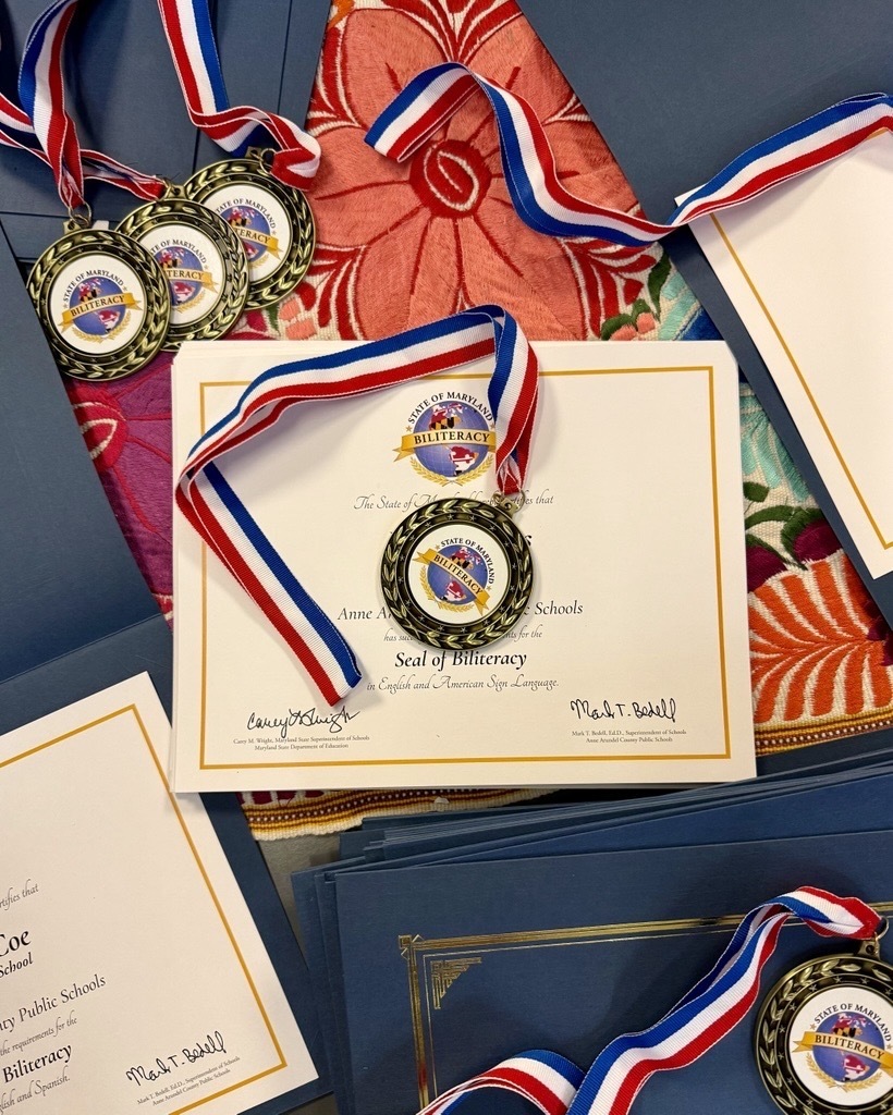 Several medals and certificates are displayed on blue folders, including one with a blue ribbon and gold medal for AACPS Maryland Seal of Biliteracy 