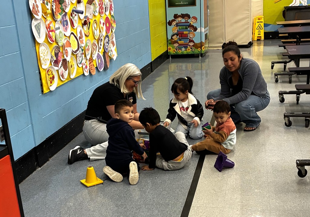 A group of children with two adult women sitting on a school floor. Some children play with toys.