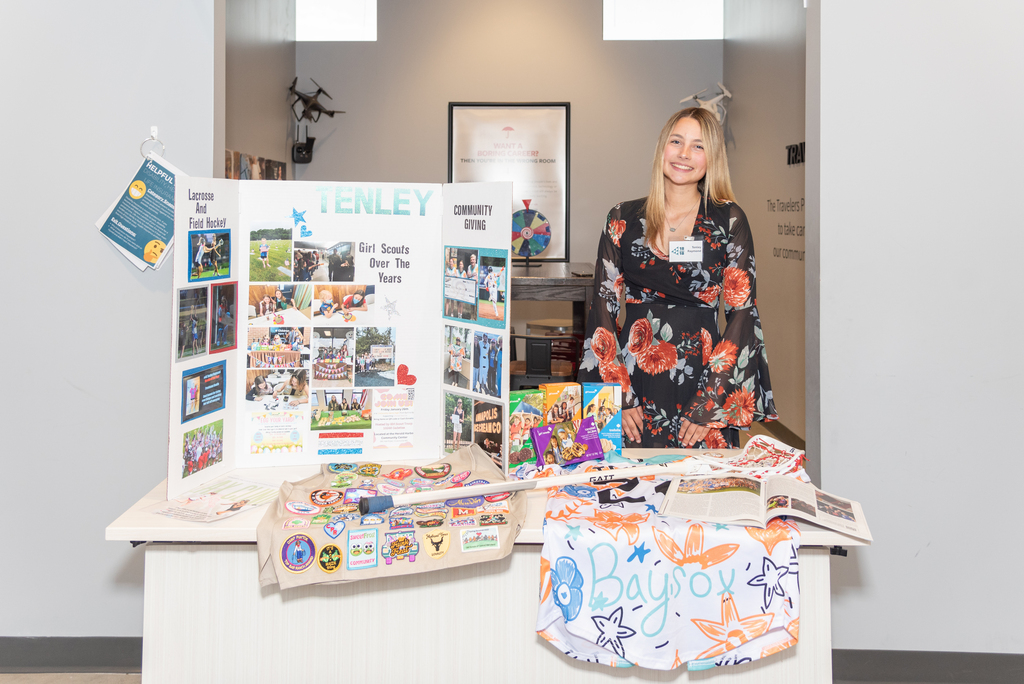 Tenley stands behind a table with a collage, a white cloth, and a banner. Items are on the table.