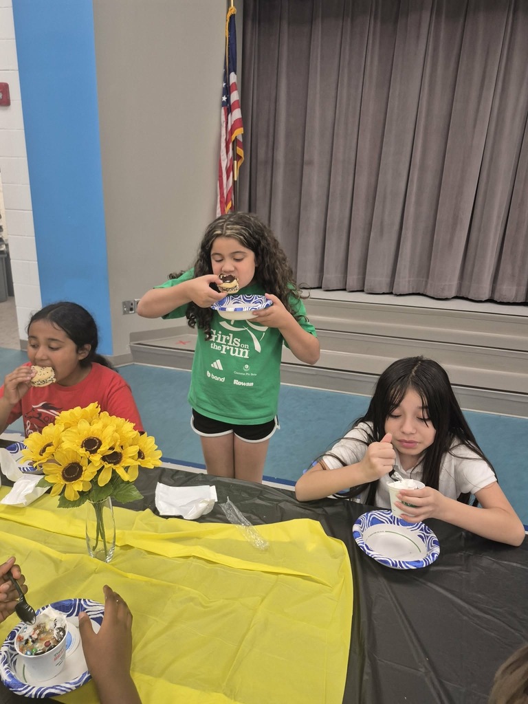 Three girls at a table eating ice cream. A sunflower vase is on the table, and an American flag hangs in the background.
