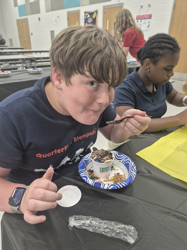 Two students, one eating ice cream, sit at a table in a cafeteria with a yellow paper.