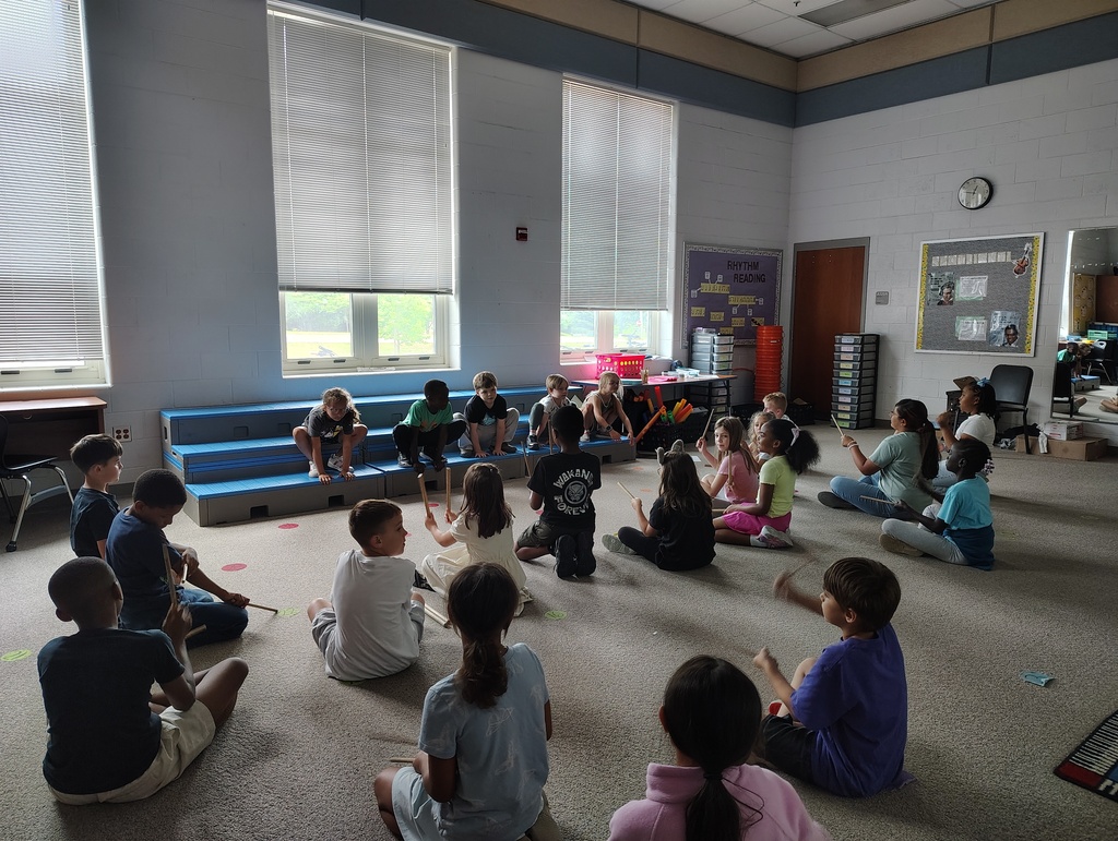 classroom of students, some on the blue risers, some on the floor with rhythm sticks
