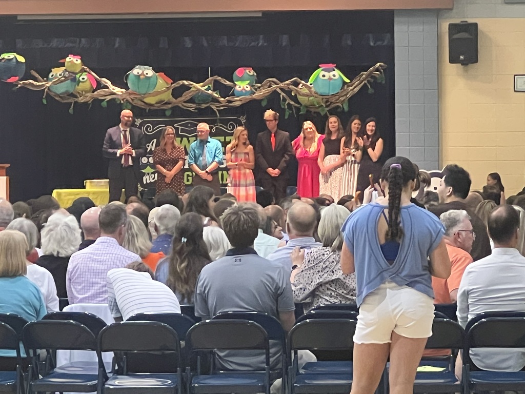 A group of people stands on stage as others watch from chairs. A yellow backdrop has bird decorations.
