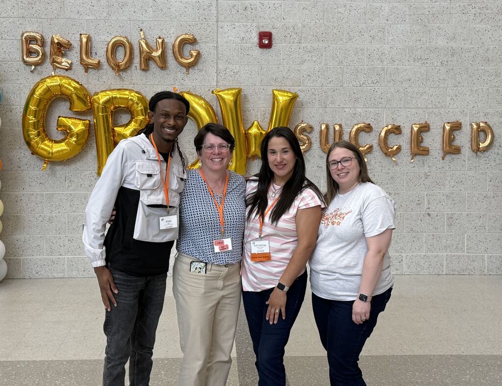 Four teachers posing in front of the county's vision of Belong, Grow, Succeed.
